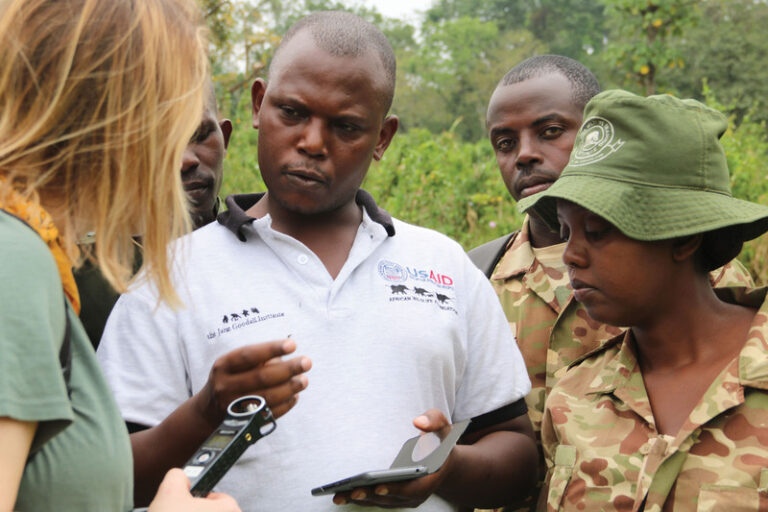 Community Forest Monitors Embrace Gender Equity in Tanzania and Uganda Photo of a masculine person with dark skin and a buzzcut haircut wearing a Jane Goodall Institute polo shirt speaking to a group of people: two masculine people with dark skin and buzzcut haircuts, one feminine person with dark skin and hair hidden under a bucket hat, and one feminine person with light skin and shoulder-length, wavy, blond hair.