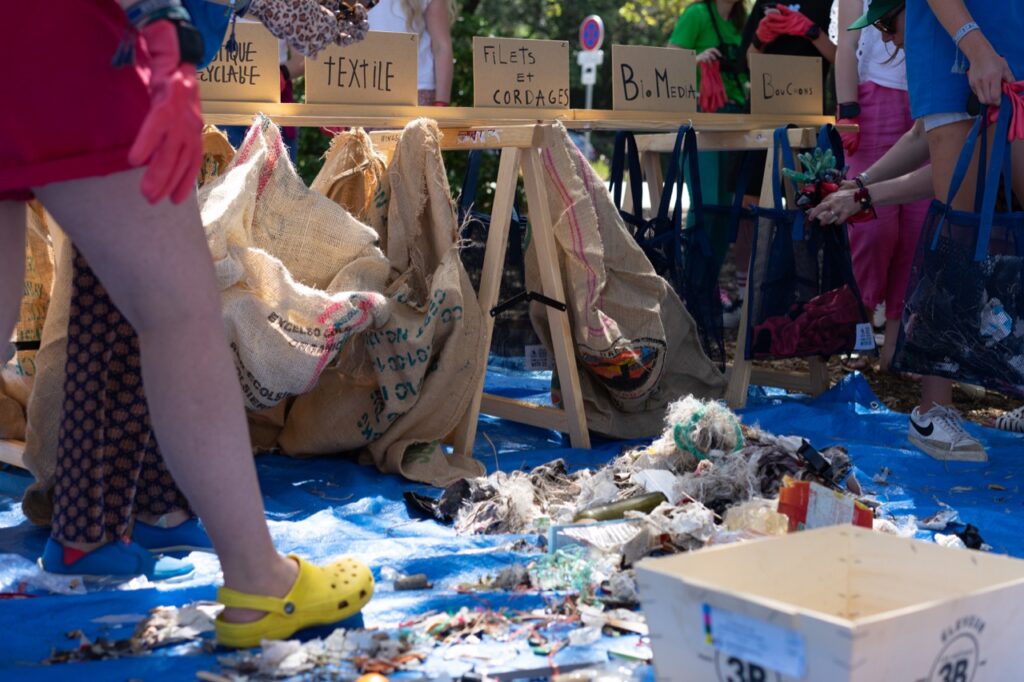 Photo of a blue tarp scattered with litter that is being sorted by people into the boxes at a table behind, each labeled in French with a different material type.