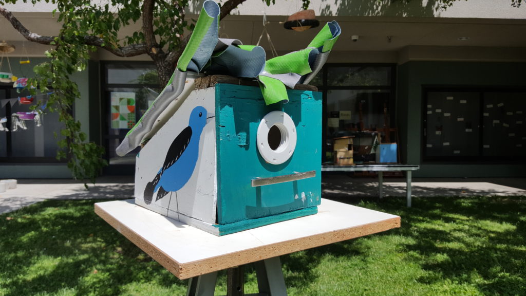 Photo of a bird house made of reused materials, and painted with a blue bird on the side.