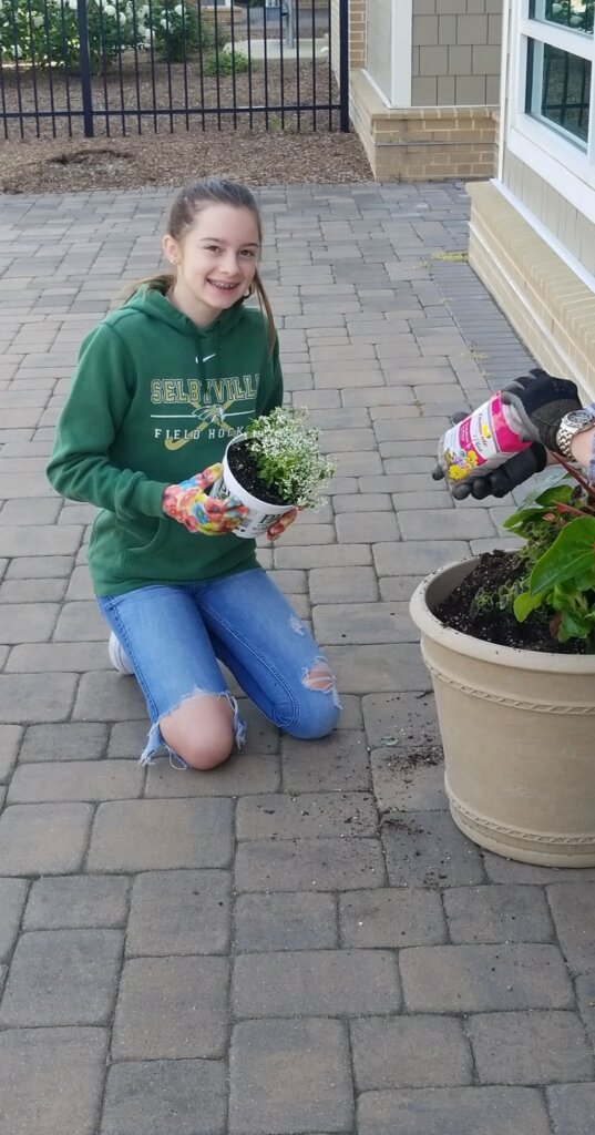 Photo of a teenager with pale skin and dark blond hair holding a potted plant that she's about to place in a larger outdoor pot.