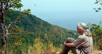 An older woman (Dr. Jane Goodall) sits on the ground, knees tucked in to her chest, with her arms clasped around her legs. She is in a beautiful outdoor setting, overlooking trees and water, and is gazing out onto the scenery. She wears her white hair tucked back into a low ponytail, a tan short-sleeved, button-down shirt and light green pants, and tan sandals.