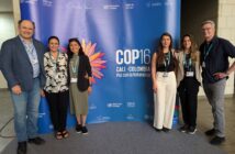 A photo of six people of varying appearances standing for a photo around a large blue sign that says, "COP16 / Cali, Colombia / Paz con la Naturaleza."