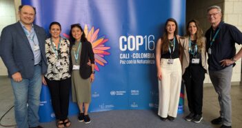 A photo of six people of varying appearances standing for a photo around a large blue sign that says, "COP16 / Cali, Colombia / Paz con la Naturaleza."