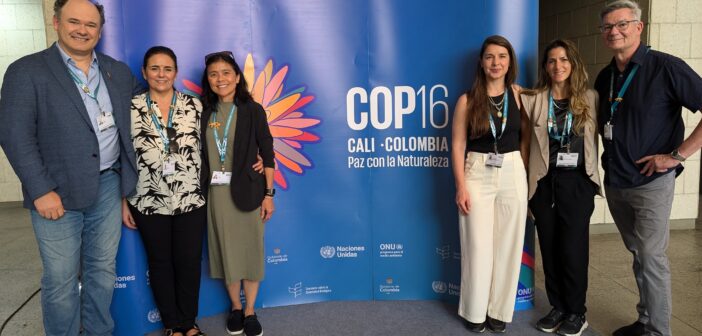 A photo of six people of varying appearances standing for a photo around a large blue sign that says, "COP16 / Cali, Colombia / Paz con la Naturaleza."