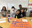 A photo of five kids standing behind a table in a classroom: a child with light brown skin and shoulder-length, straight, black hair; a child with light brown skin and wearing a beanie; a child with light brown skin and short, straight, black hair; a child with light brown skin and long, wavy, black hair; a child with dark skin and short, curly, brown hair. On the table in front of them is a large poster that says, "Donate to help animals / Thank you!"
