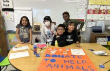 A photo of five kids standing behind a table in a classroom: a child with light brown skin and shoulder-length, straight, black hair; a child with light brown skin and wearing a beanie; a child with light brown skin and short, straight, black hair; a child with light brown skin and long, wavy, black hair; a child with dark skin and short, curly, brown hair. On the table in front of them is a large poster that says, "Donate to help animals / Thank you!"