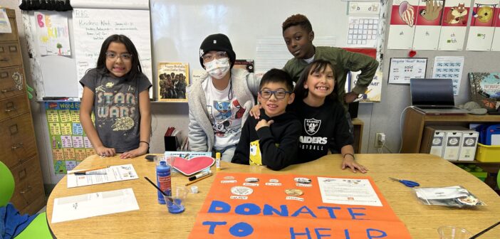 A photo of five kids standing behind a table in a classroom: a child with light brown skin and shoulder-length, straight, black hair; a child with light brown skin and wearing a beanie; a child with light brown skin and short, straight, black hair; a child with light brown skin and long, wavy, black hair; a child with dark skin and short, curly, brown hair. On the table in front of them is a large poster that says, "Donate to help animals / Thank you!"